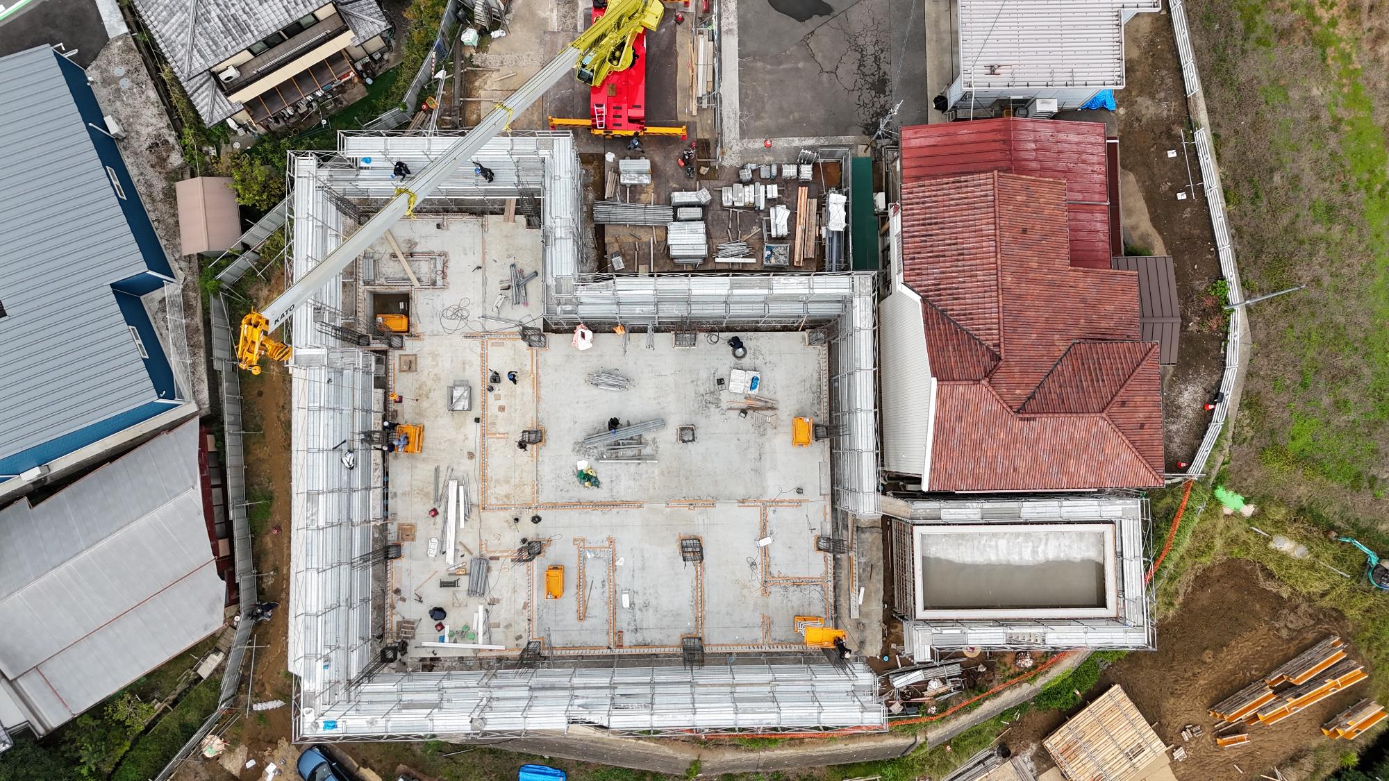令和7年10月3日時点の消防署富来田分署建設現場を真上から撮影した写真です。建設中の建物は大きな四角形の形状をしており、周囲を足場が取り囲んでいます。建物内部では1階部分のコンクリート床が施工され、柱や壁となる鉄筋が各所で立ち上がっています。中央部や周辺部には将来の部屋や廊下の配置を示すように型枠や墨出し線が設けられ、建物の内部構造が形になり始めている様子が確認できます。現場内では複数の作業員が作業を行い、大型クレーンのブームが建物上空まで伸びています。建物の右側には既存の消防施設が隣接し、その周囲には資材置場や作業スペースが設けられています。この写真からは、基礎工事を終え、建物の骨組みとなる柱や壁の施工が本格的に進められている段階であることが分かります。