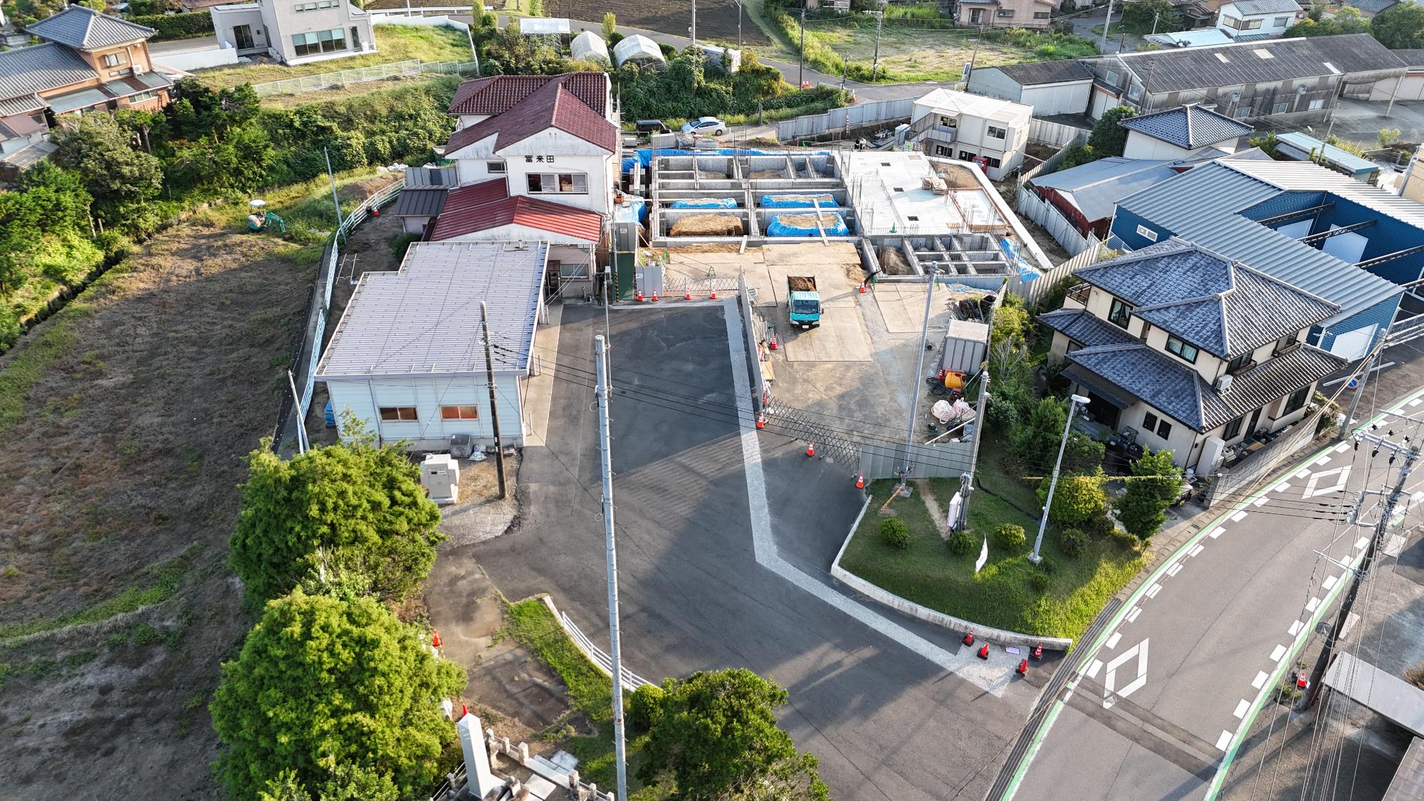 令和7年9月10日時点の消防署富来田分署建設現場を斜め上空から撮影した写真です。写真中央奥では新庁舎の建設に向けた基礎工事が進められており、格子状に配置されたコンクリートの基礎梁が建物の輪郭を形作っています。一部の区画には土が残され、青色のシートで覆われた箇所も見られます。建設予定地の周囲には仮囲いや安全柵が設置され、工事車両や資材が配置されています。左側には既存の消防署庁舎があり、「富来田」の表示が確認できます。また、周辺には住宅や農地が広がり、地域に囲まれた環境の中で建設工事が進められている様子が分かります。この時点では建物本体の着工初期段階であり、建物を支える基礎構造の施工が進められている状況です。
