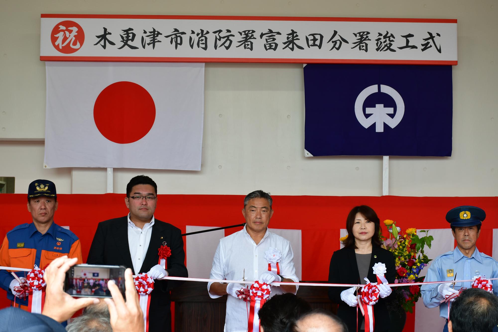 木更津市消防署富来田分署竣工式の様子を撮影した写真です。会場正面には紅白幕が張られ、その上には「祝木更津市消防署富来田分署竣工式」と書かれた看板が掲げられています。看板の下には日本国旗と木更津市旗が飾られています。写真中央ではテープカットが行われており、参加者が白い手袋を着用して赤白のリボンが付いたテープをはさみで切る瞬間が写されています。向かって左から消防団山田副団長、草刈市議会議長、渡辺市長、廣橋副市長、宗政消防長の5名が横一列に並んでいます。消防関係者は制服を着用し、市の関係者は式典用の胸章を付けています。参列者が見守る中、新しい富来田分署の完成を祝い、施設の門出を記念する厳かな式典の様子が表現されています。