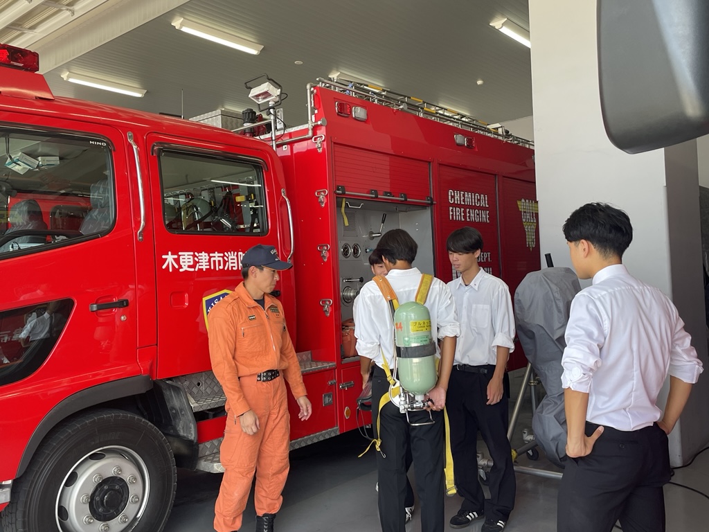 消防車両の資機材について消防職員が説明をし、説明会参加者が実際に空気ボンベを背負っている様子。