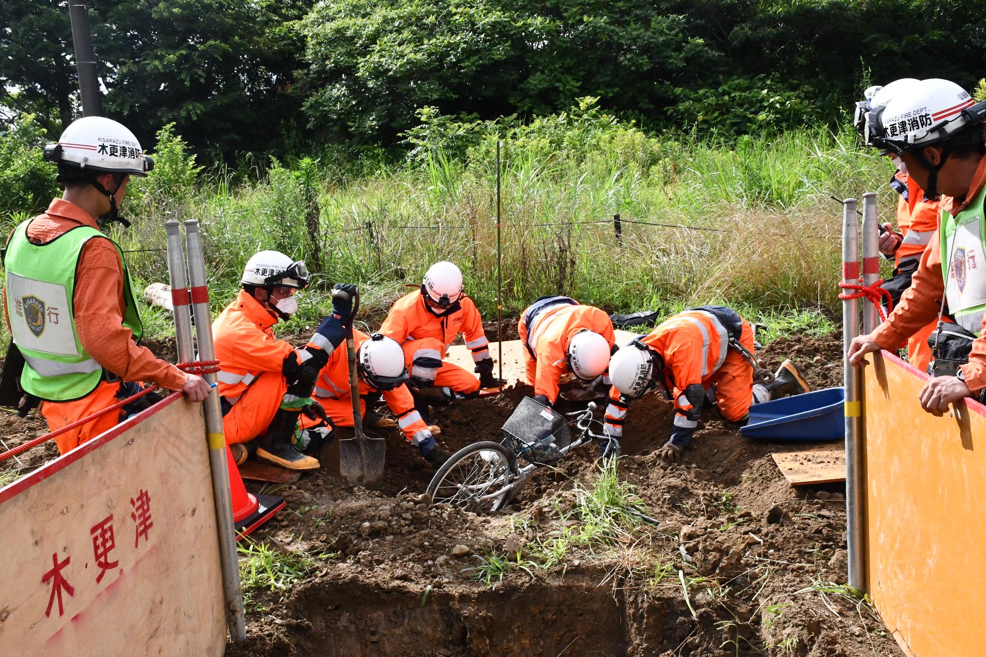 青空の下、土砂災害対応訓練会場で土砂に埋まった自転車の周りを要救助者人形がいる可能性があることから、大勢の消防職員が手掘りで掘っている様子。