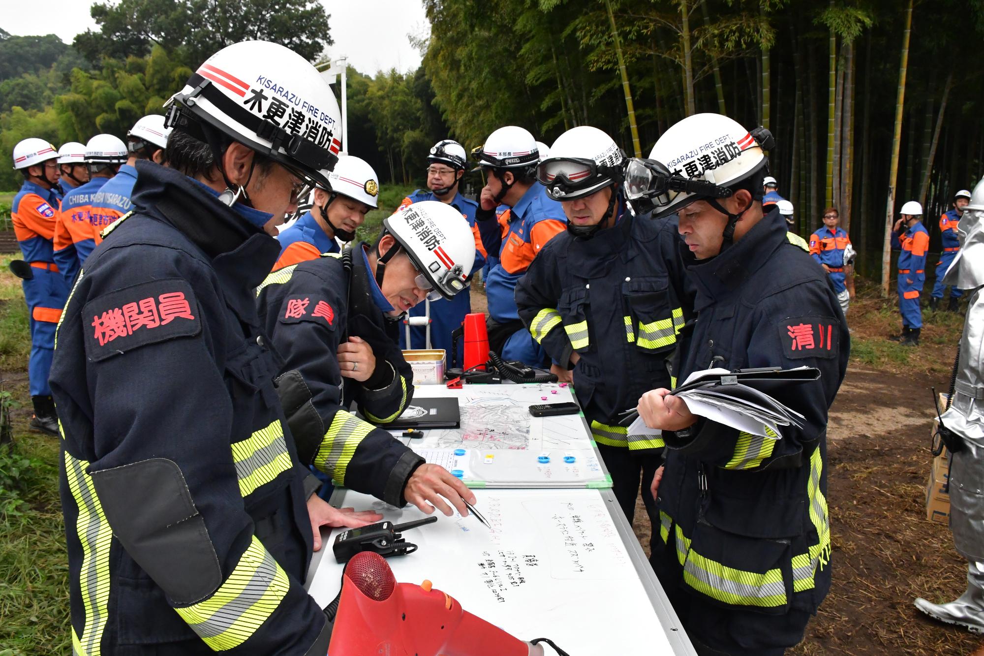 青空の下、林野火災対応訓練の会場で設定した現場指揮本部において、消防職団員2名に対し、現場指揮の消防職員2名が机上に置かれた作戦ボードを見ながら消防職員に消火作業指示など現場指揮している様子。