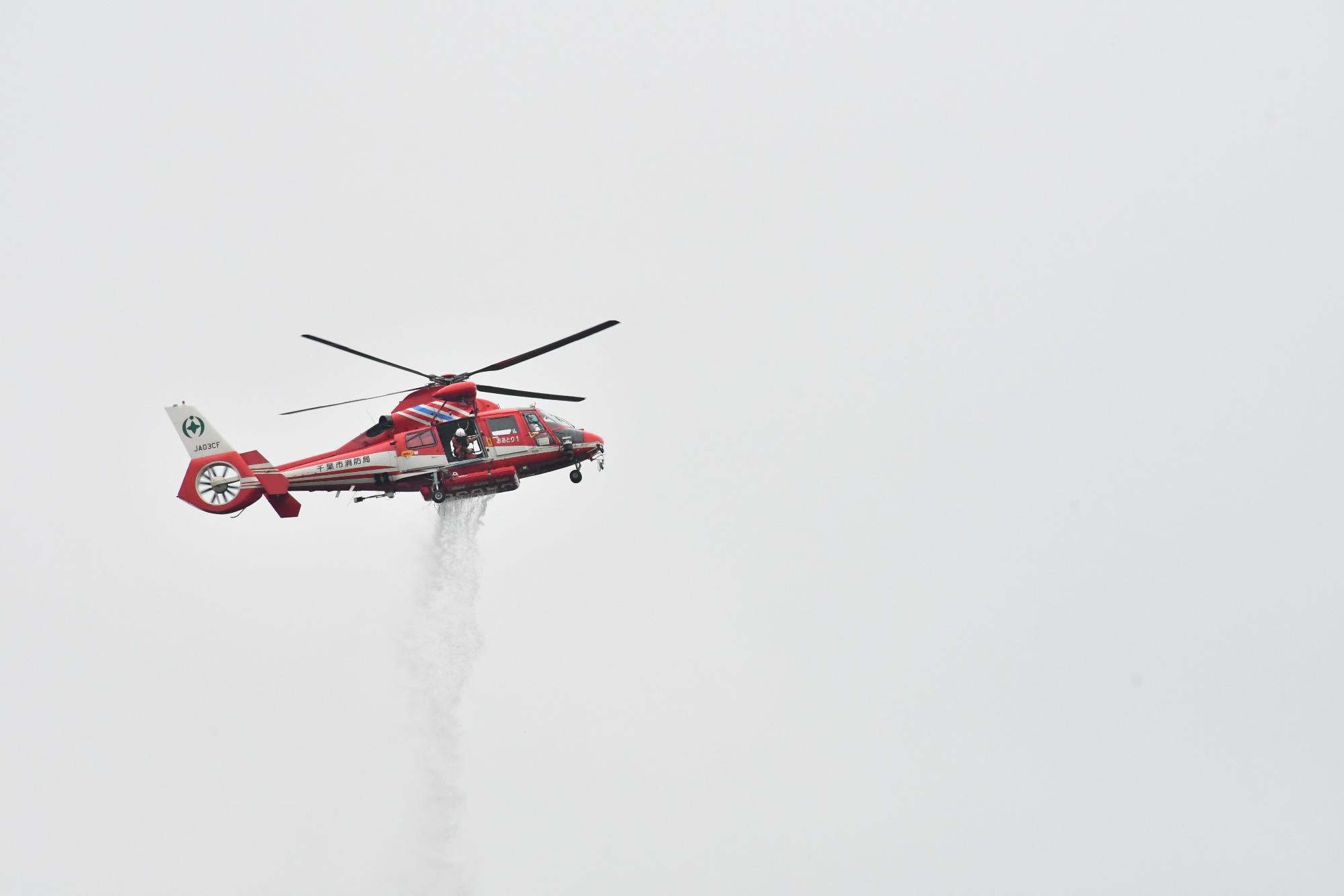 曇り空の下、林野火災が想定される地点に消防ヘリが空中消火のためヘリの下部から消火用水を放水している様子