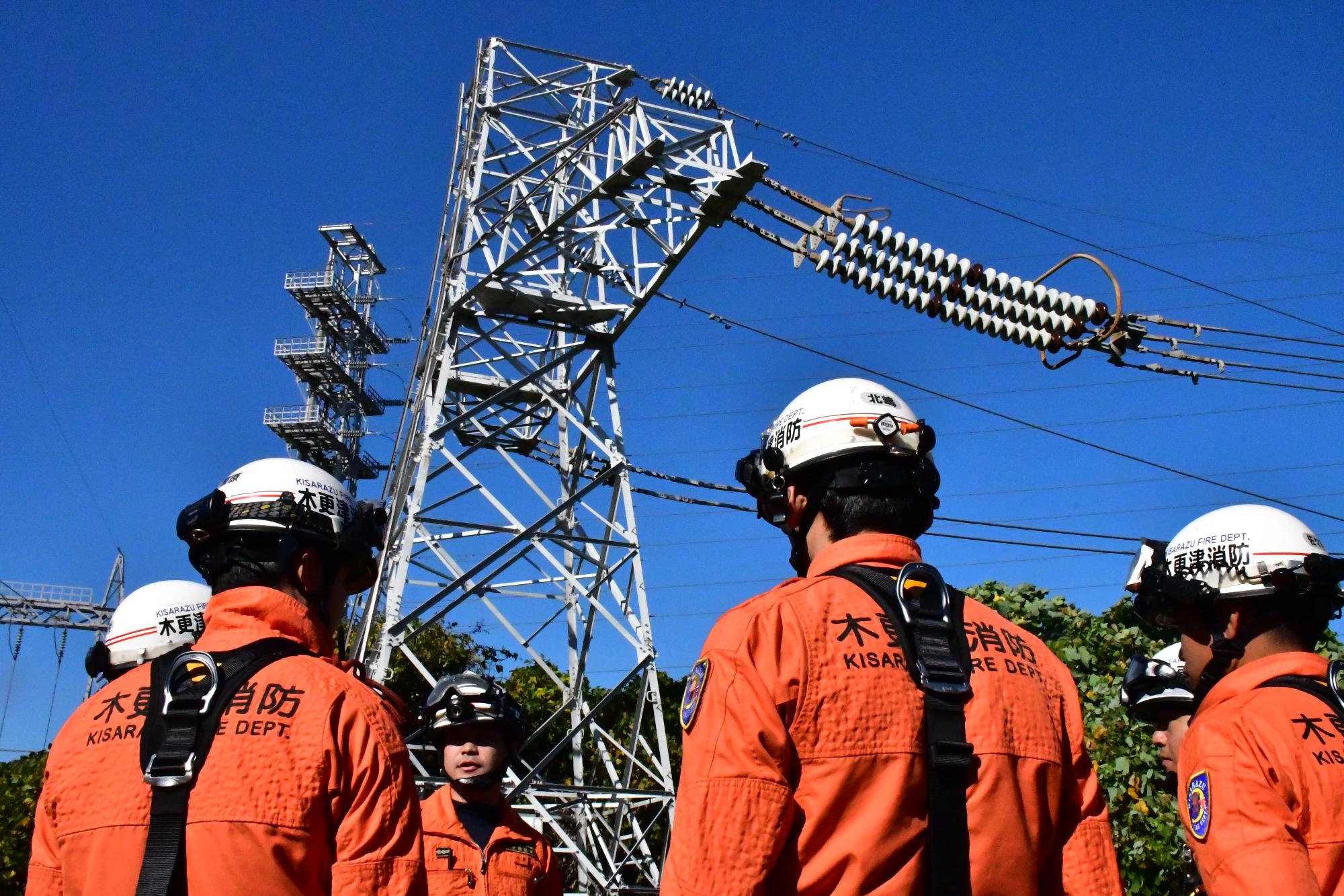青空の下、送電鉄塔の上部で宙吊りになった訓練人形を、ヘルメットを被りオレンジ色の救助服を着用した5名の救助隊員がこれから救出しようと隊員間での指示内容の確認などしている様子