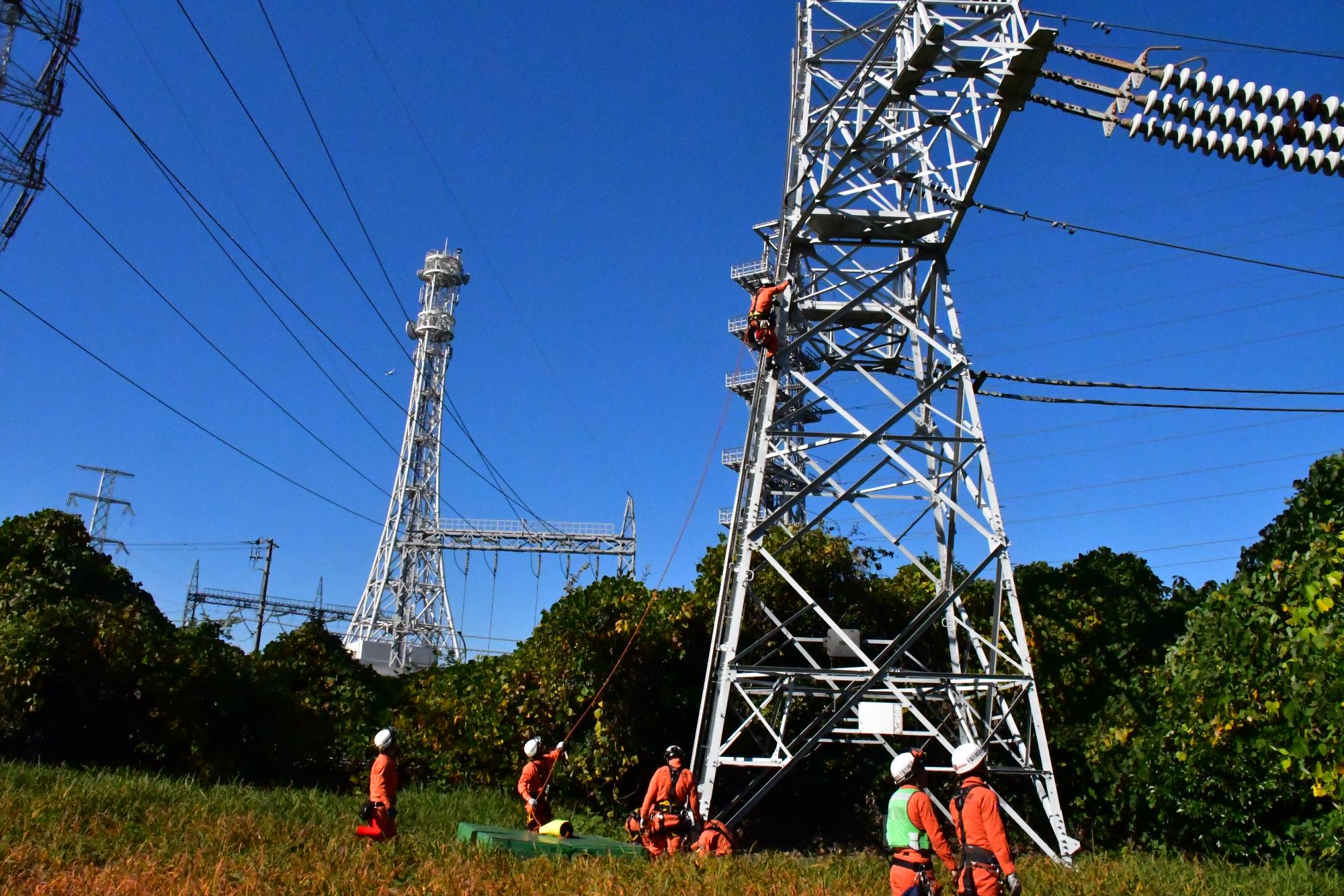 ヘルメットを被りオレンジ色の救助服を着用した救助隊員が、鉄塔上部にいる傷病者とみたてた訓練人形を救出するため、鉄塔を登っている様子