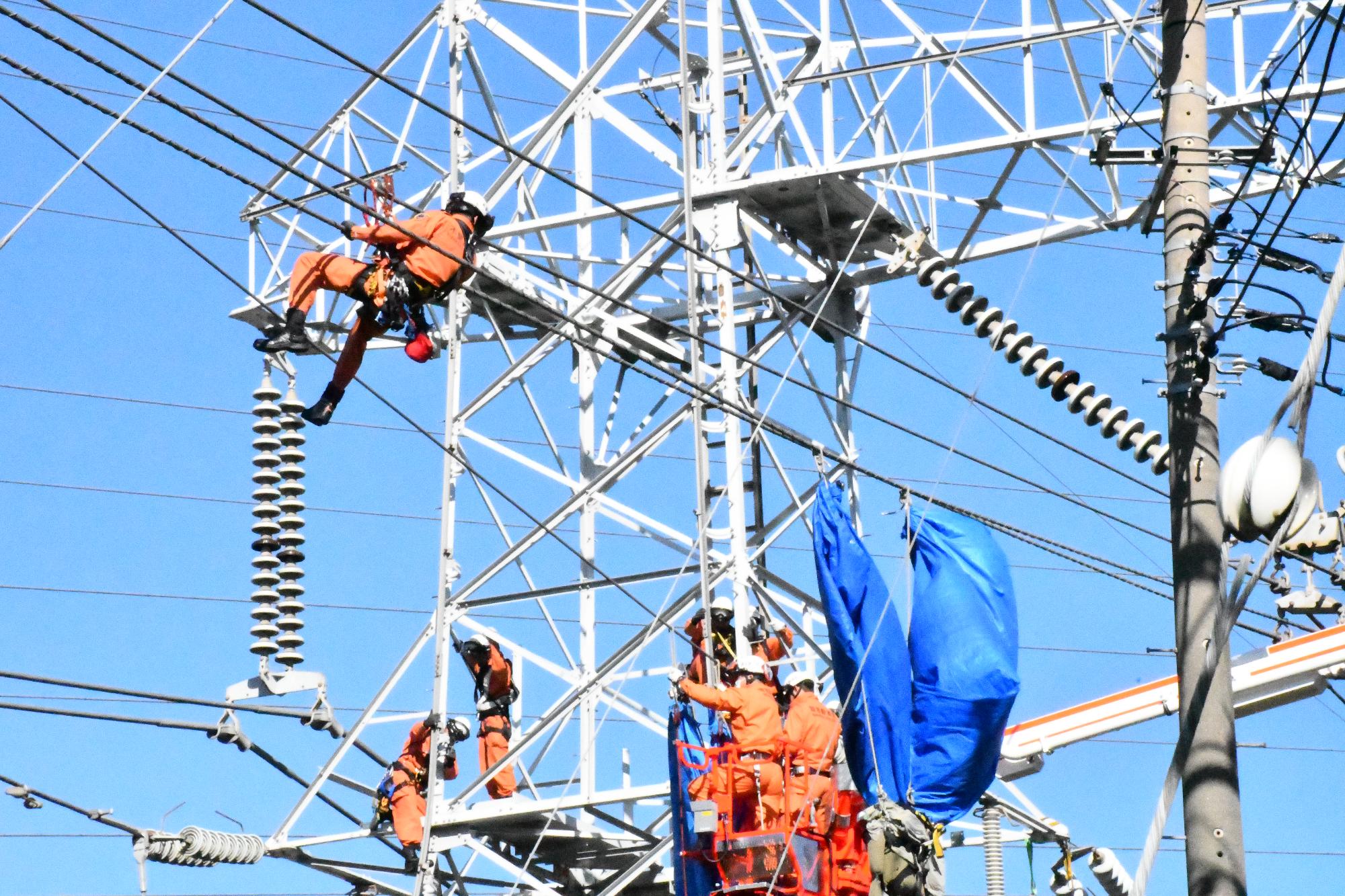 青空の下、ヘルメットを被りオレンジ色の救助服を着用した1名の救助隊員が鉄塔上部で要救助者とみたてた訓練人形を救出するため送電線を渡っている様子
