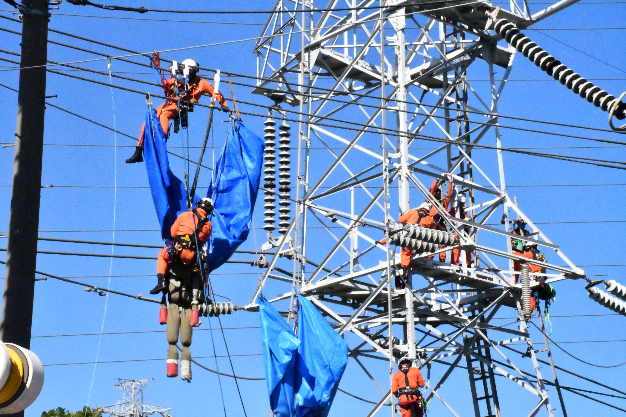 青空の下、オレンジ色の救助服を着用した救助隊員2名が送電線に宙吊りになっている訓練人形を救出している様子