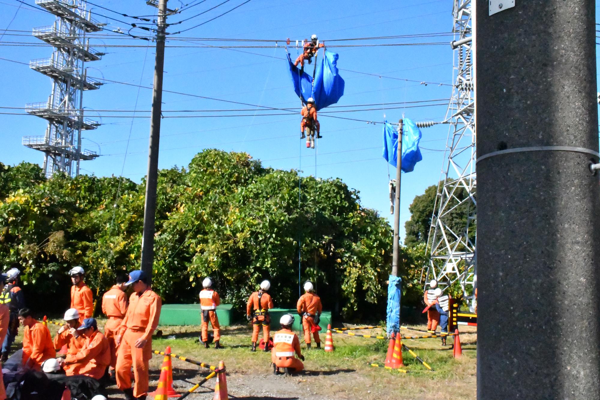 青空の下、オレンジ色の救助服を着用した救助隊員2名が電線にぶら下がっている訓練用人形を救出しようとしているところを少し離れたところから撮影した様子