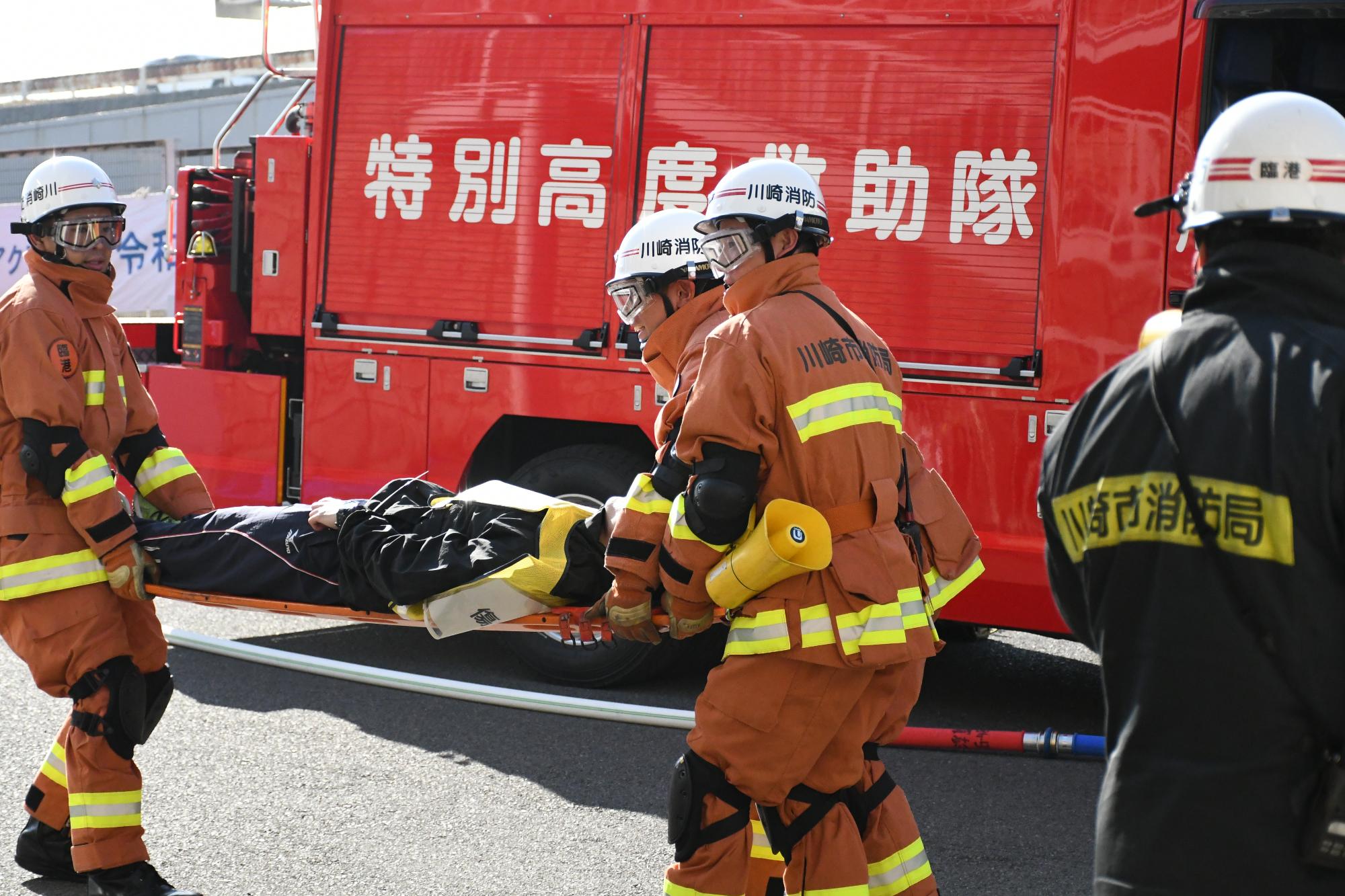青空の下、救助隊3名が訓練事故車両から救出された傷病者を重症度・緊急度別に分けるスペースに搬送している様子