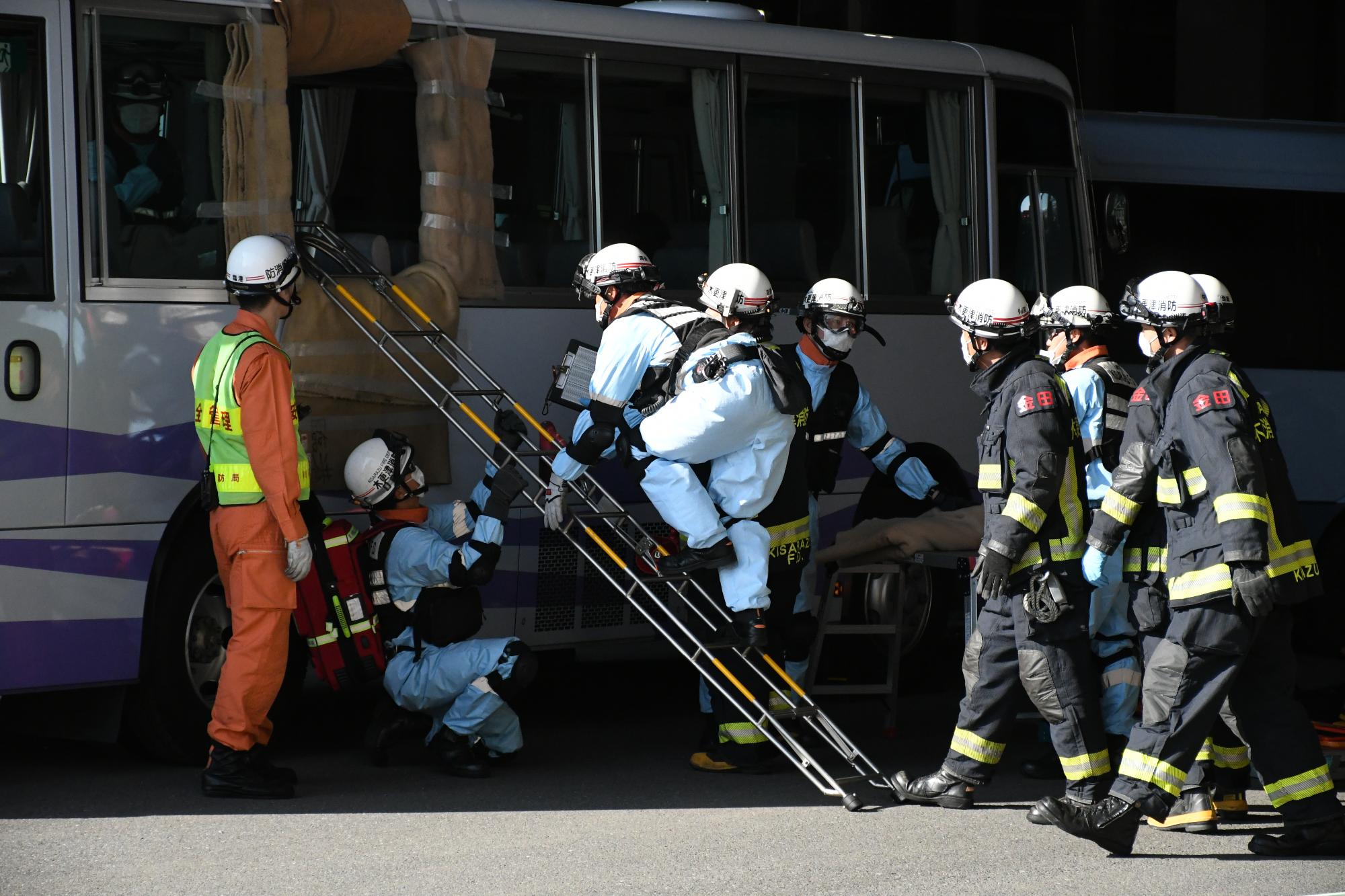 青空の下、訓練事故車両の大型バス内にいる要救助者を救出するため、バスの窓にかけた梯子から消防隊が救出するためにバス内に進入しようとしている様子