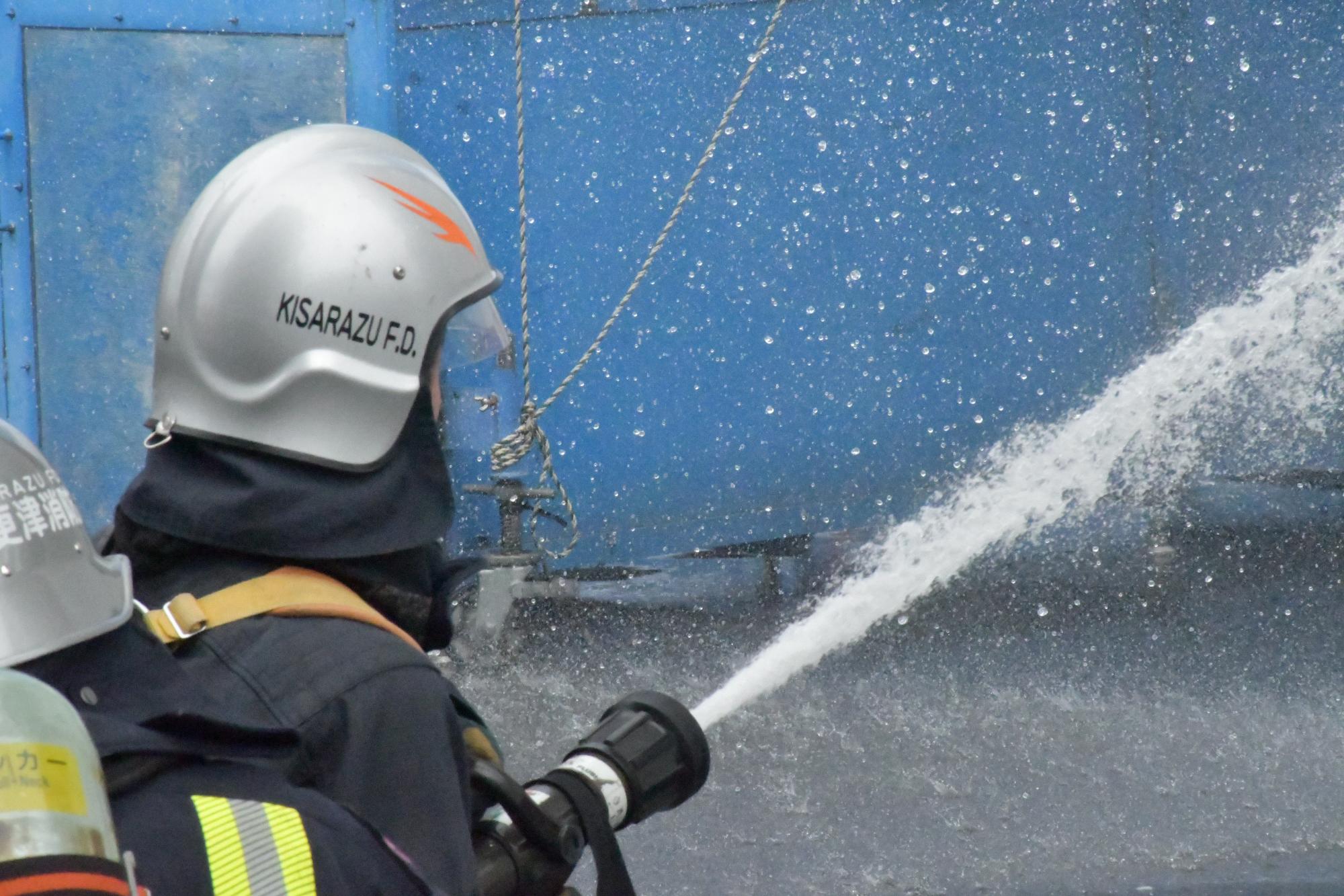 防火衣を着装し空気呼吸器を背負った1名の新規採用消防職員が、訓練塔に向けて放水を行っている様子の写真