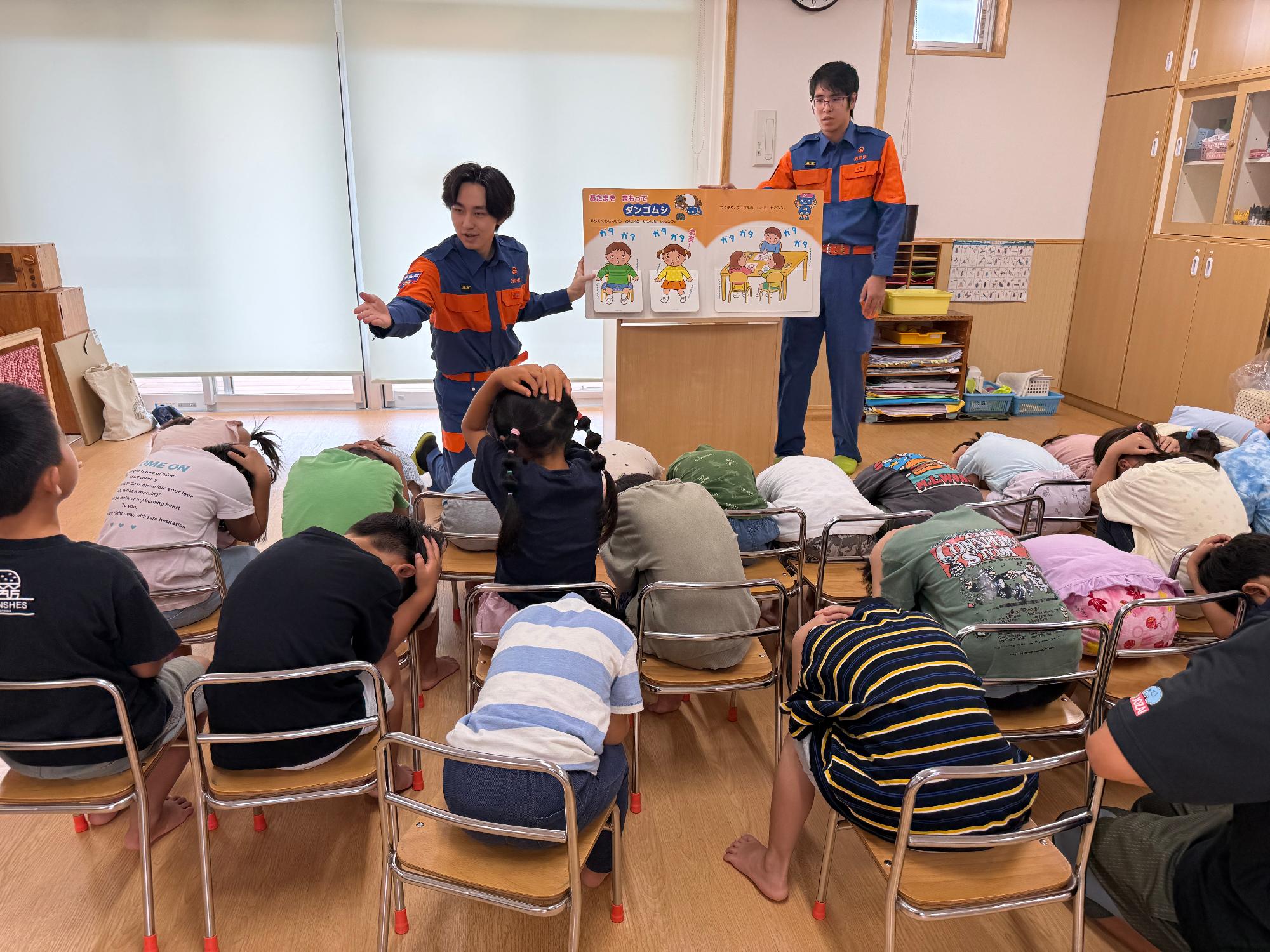 保育園の教室で消防団機能別分団学生部2名が園児たちに対して防災啓発用の紙芝居を読み聞かせている様子