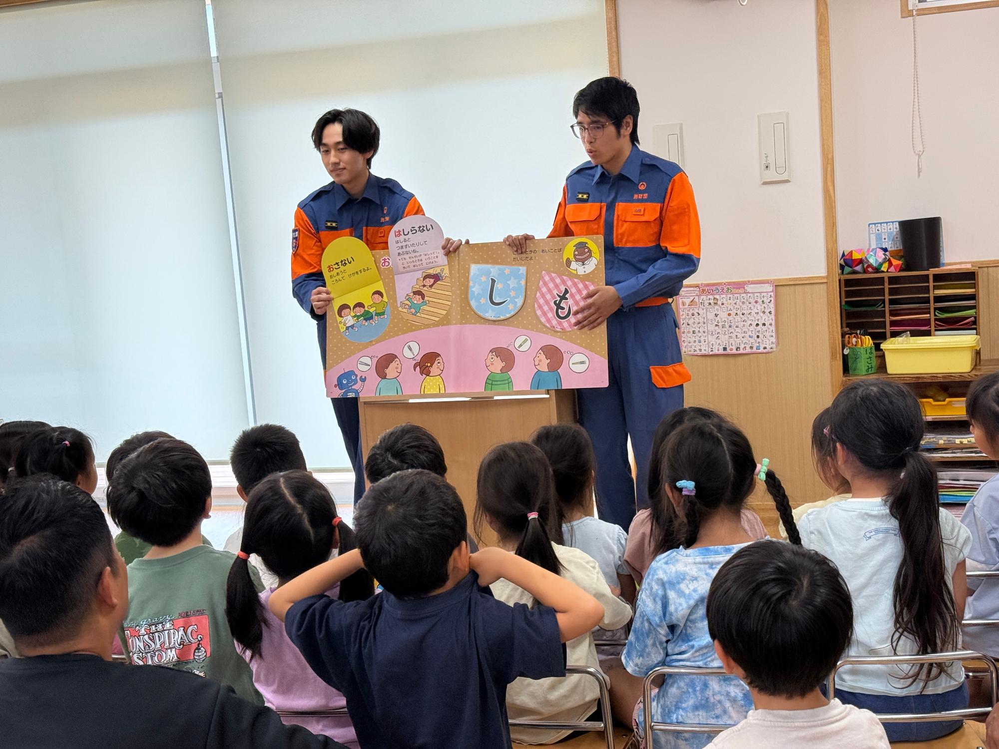 保育園の教室で消防団機能別分団学生部2名が園児たちに対して防災啓発用の紙芝居を読み聞かせている様子