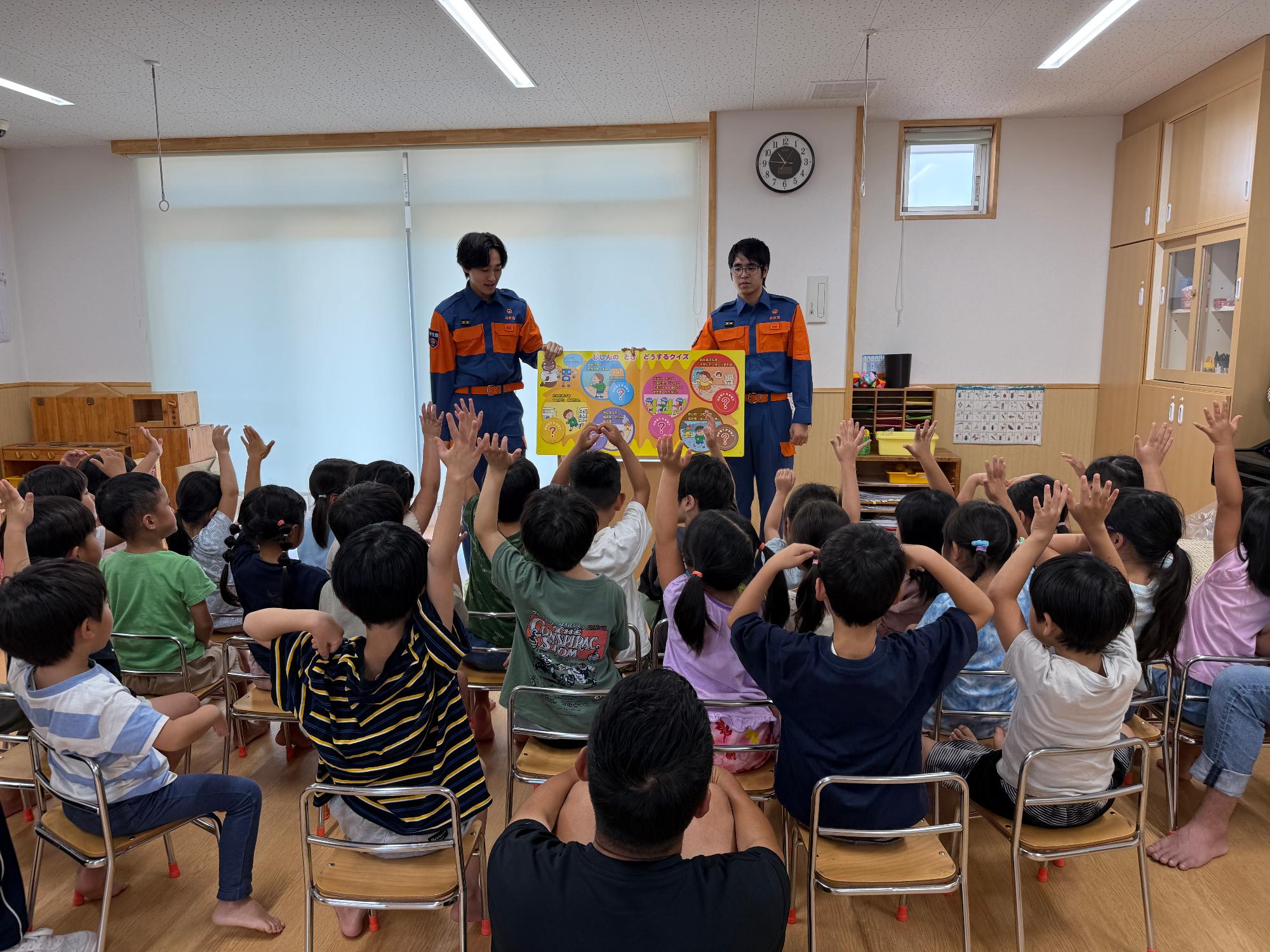保育園の教室で消防団機能別分団学生部2名が園児たちに対して防災啓発用の紙芝居を読み聞かせている様子