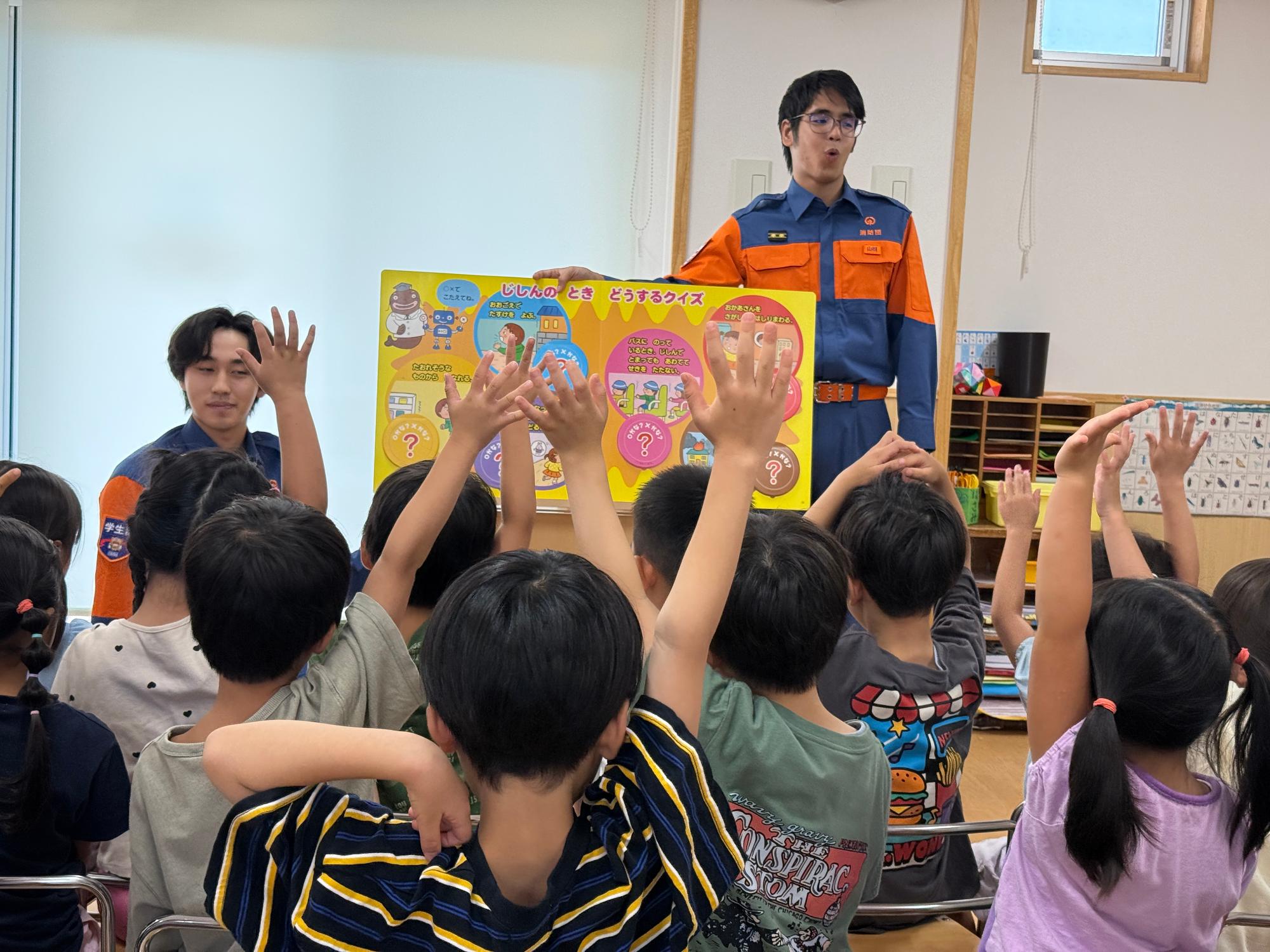 保育園の教室で消防団機能別分団学生部2名が園児たちに対して防災啓発用の紙芝居を読み聞かせている様子