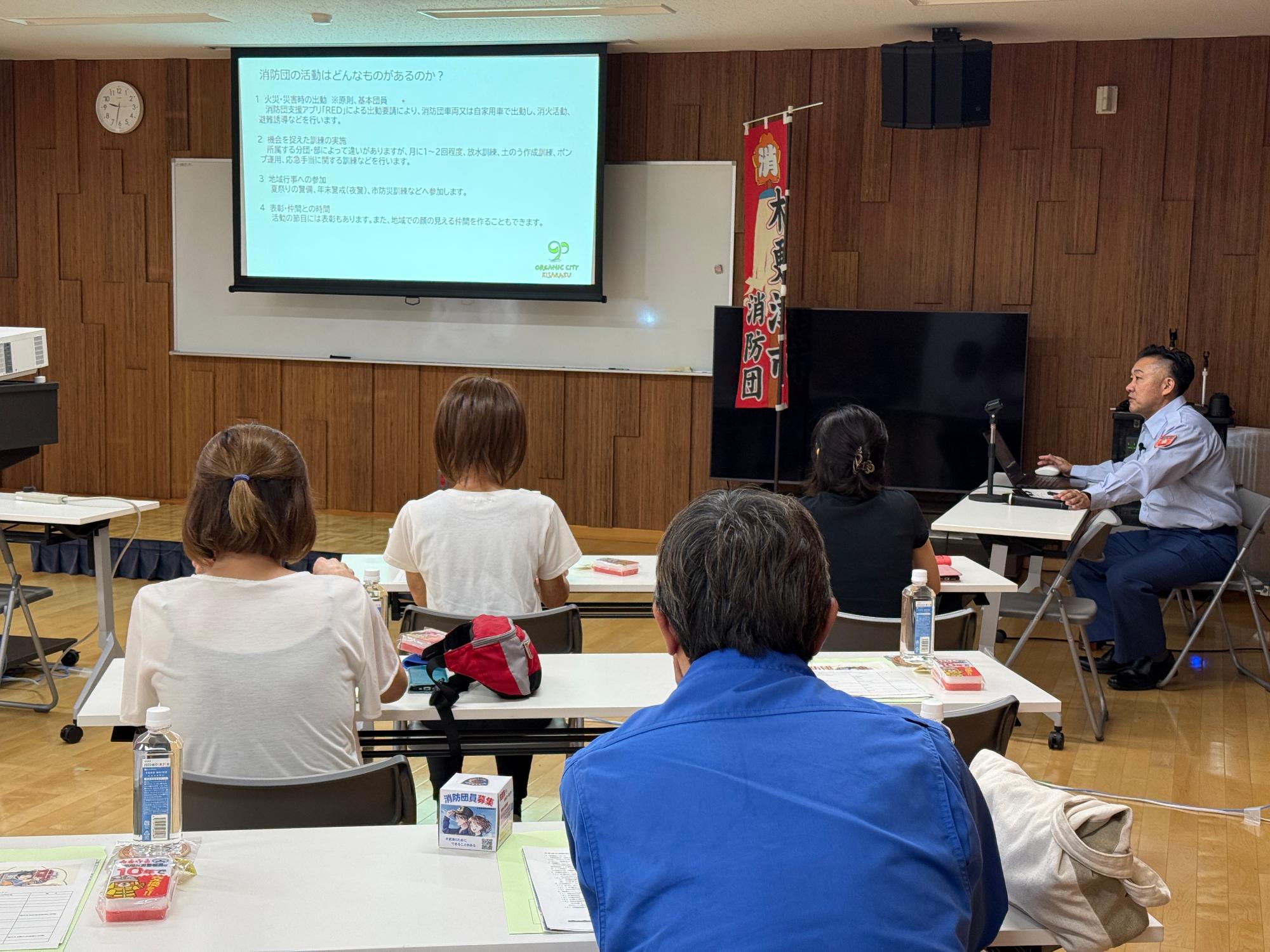 消防本部庁舎内の会議室において、消防団の役割などについて講話をしている職員と参加者を写している様子