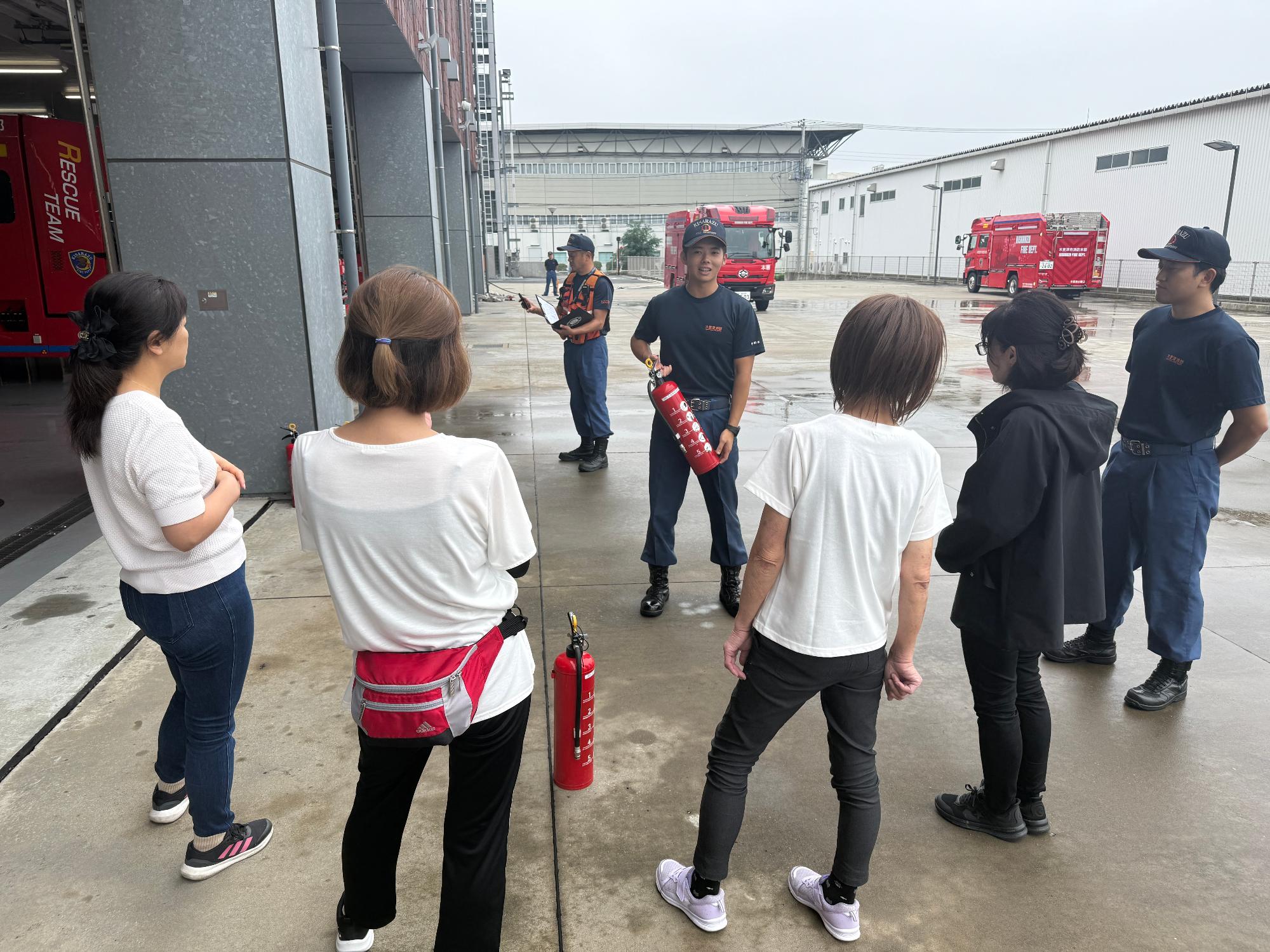 消防本部敷地内車庫前において、体験会参加者4名が消火器の取り扱い説明を受けている様子。