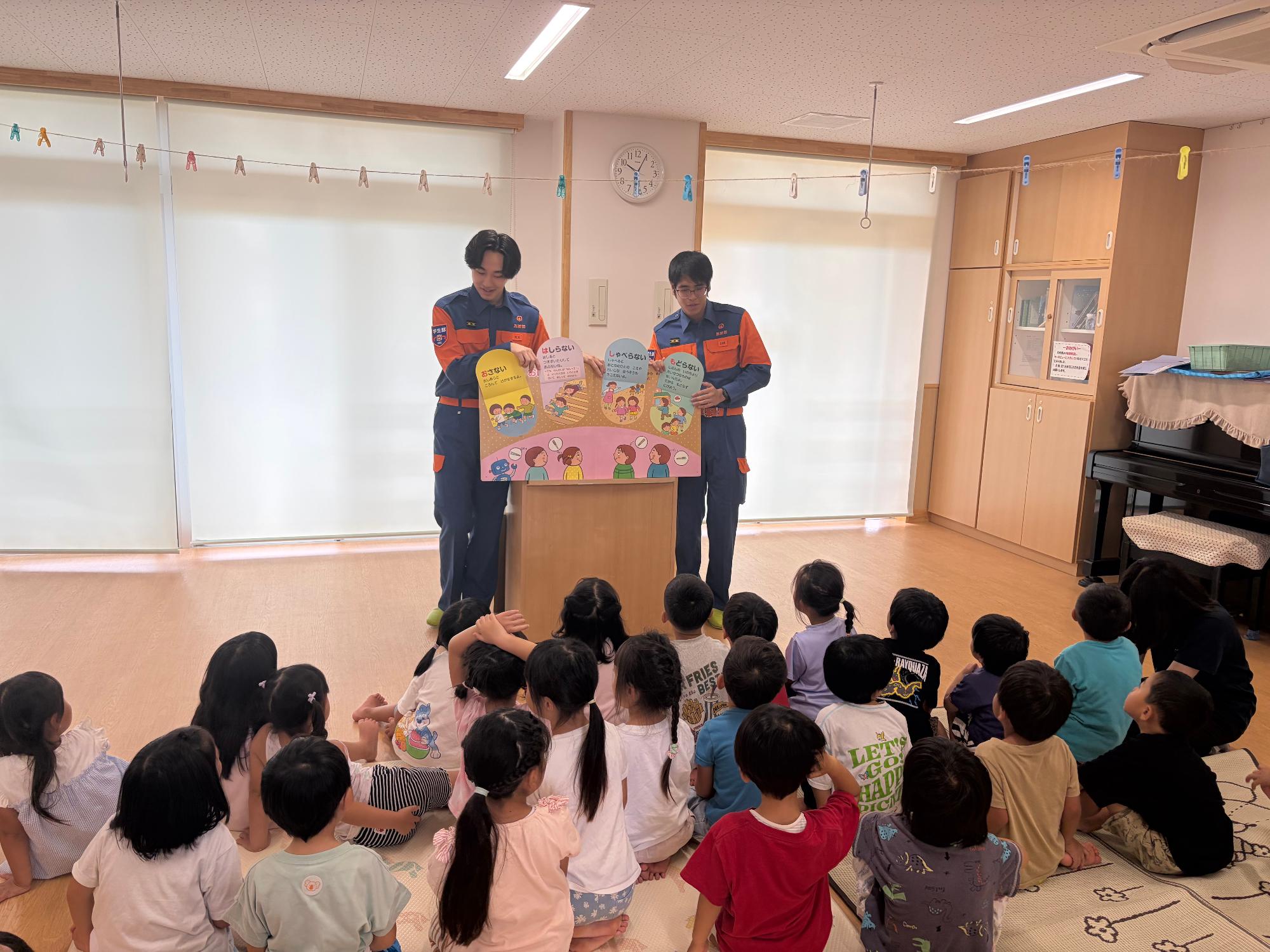 保育園の教室で消防団機能別分団学生部2名が園児たちに対して防災啓発用の紙芝居を読み聞かせている様子