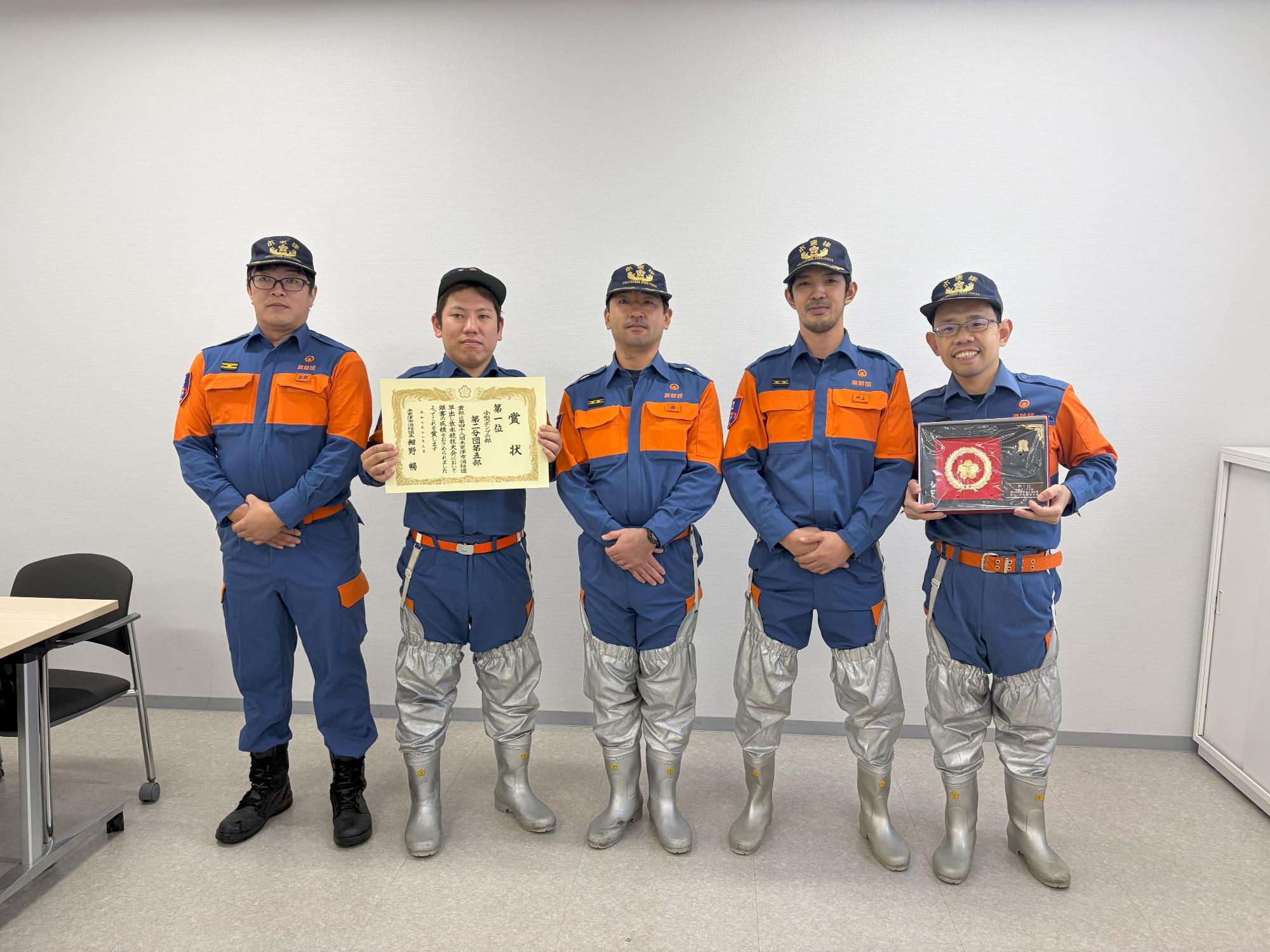 消防団員5名が、左から2番目の方が賞状、右から1番目の方が盾を持ちそれ以外は手を前にくみ横に並んで立っている記念写真