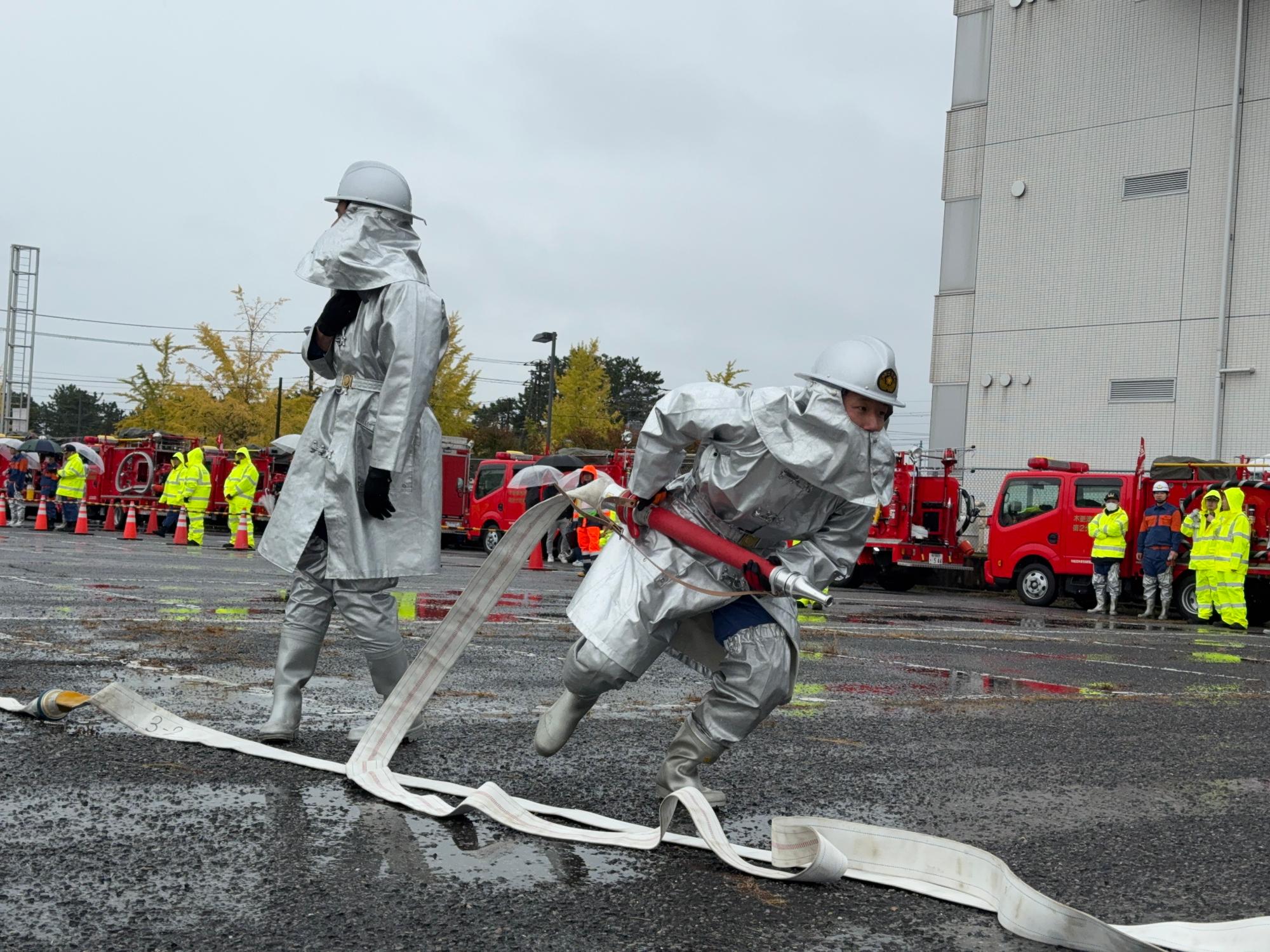 消防団員が放水の準備のためホースを管そうに結合し、放水位置へ向かうところを撮影した様子