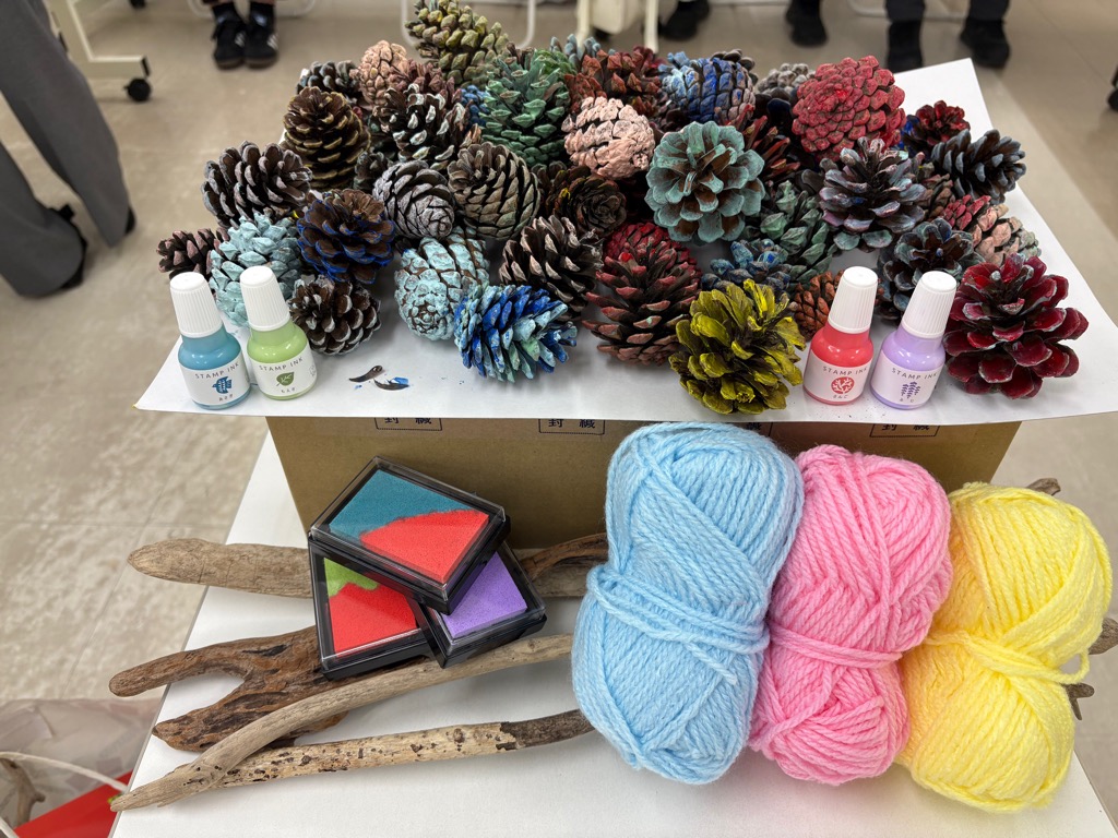 カラフルにデザインされた松ぼっくりと毛糸、流木が机の上にあります。