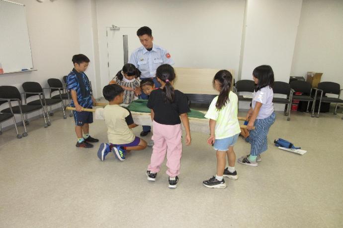 緑色の人形を子供たちが毛布と竹で作成した担架を使って持ち上げている様子