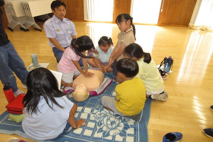 子供が人形に胸骨圧迫を実施しており、他の子供が見学している様子