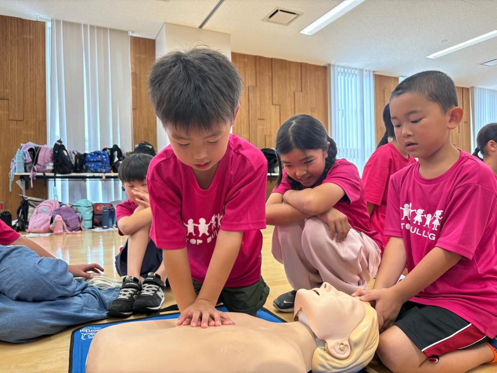 子供が人形に胸骨圧迫を実施しており、他の子供が見学している様子