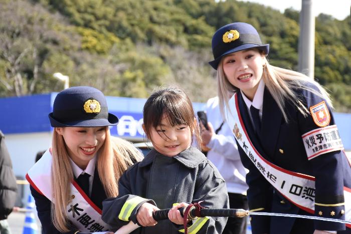 消防広場でこども用消防車による放水体験をしている様子（左からC-Style八剱咲羅、参加児童、C-Style潮干狩鯏）