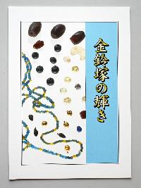 『金鈴塚の輝き』という冊子の表紙のカラー写真。表紙には金鈴塚古墳から出土した小さな金色の鈴、白色半透明の瑪瑙の勾玉、青色や黄色のガラス小玉のネックレスなどが写っています。