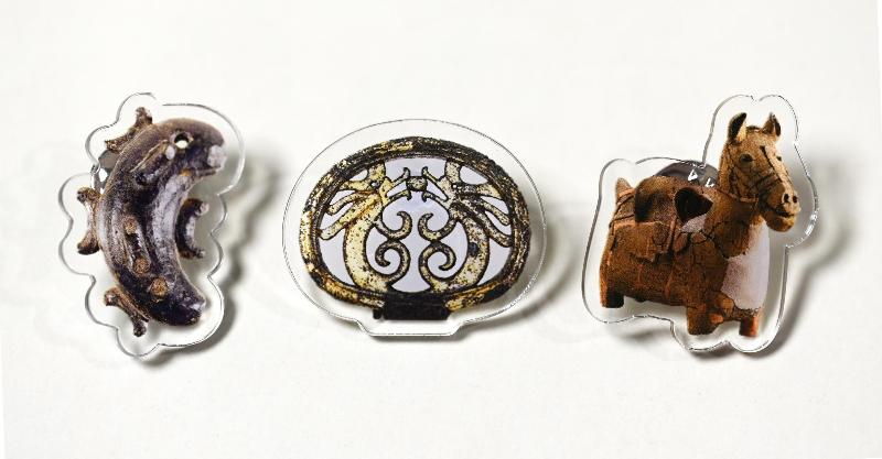 木更津市郷土博物館金のすずのアクリルピンバッチ3点が並んだカラー写真。左から順に、マミヤク遺跡から出土した灰色の子持勾玉、金鈴塚古墳から出土した金色の双龍環頭大刀の柄頭、畑沢埴輪窯跡から出土した茶色の馬形埴輪の写真を印刷したピンバッチが並んでいます。
