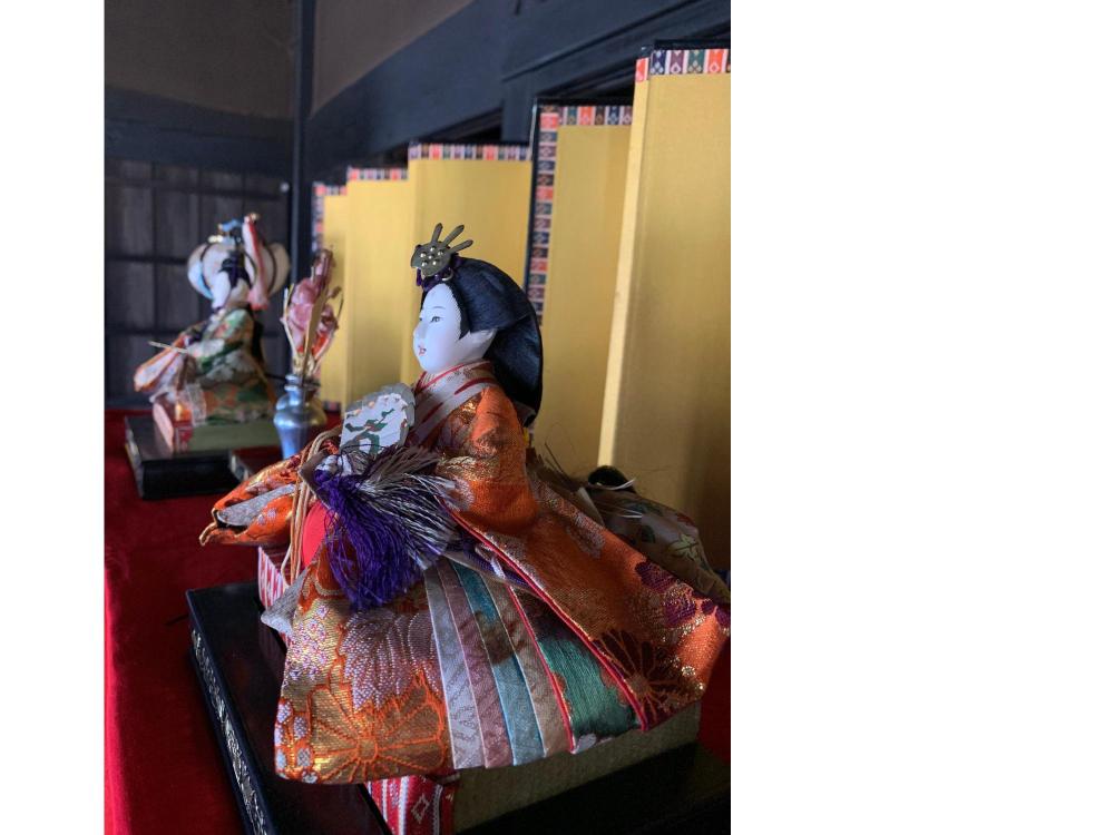 旧安西家住宅内に飾られた段飾りのお雛様を斜め右からアップで撮影したカラー写真。 オレンジ色の着物を着たお雛様は、お顔の正面から光を受けて、微笑んでいるように見えます。