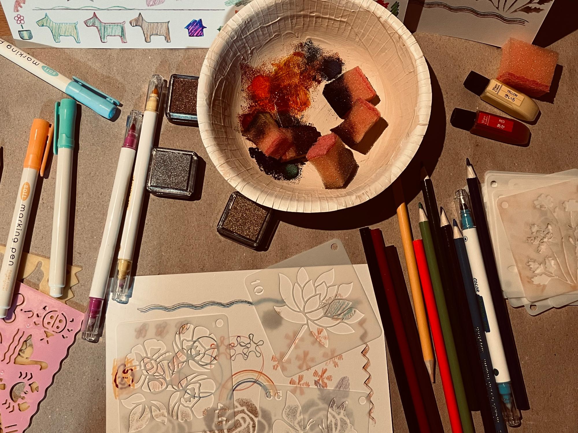 ステンシル版画を作っている机を写したカラー写真です。ステンシルシート、スタンプインク、インクのついたスポンジ、カラーペンや色鉛筆などが複数あります。