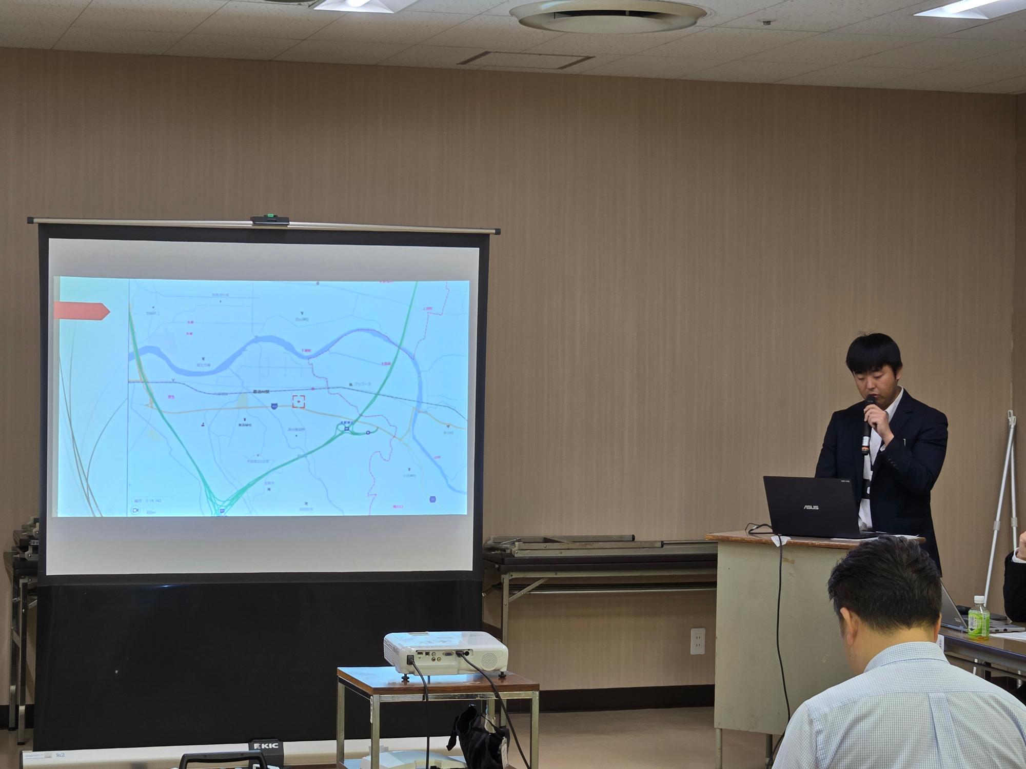 住民説明会の様子です。危機管理課の職員が、該当地区の地域特性や場所についての詳細をスクリーンに投影しながら説明しています。