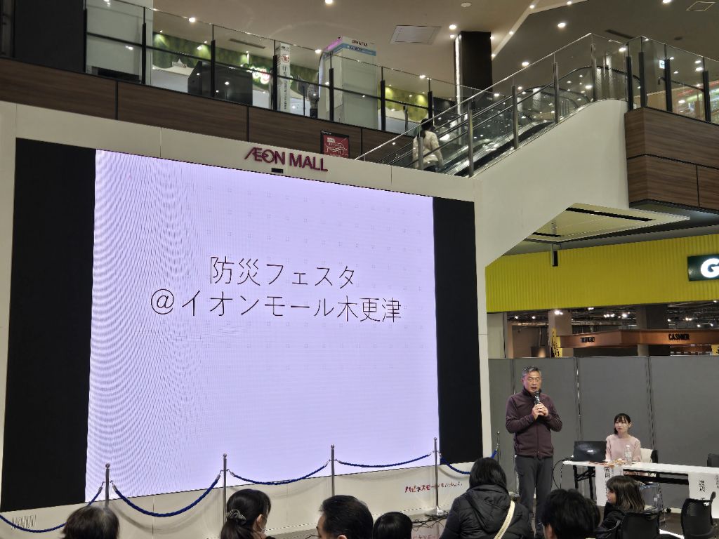 イオンモール木更津サウスアトリウムの大型スクリーンの前で参加者の皆様に挨拶を行う渡辺芳邦木更津市長の写真です。