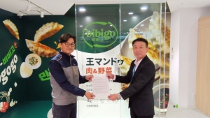 CJ FOODS JAPAN株式会社様が製造している商品が描かれた事務所の前で、締結した協定書を千葉工場長のイ・チョルソン様（左側）と木更津市総務部前田部参事（右側）で持って撮影した記念写真。