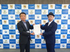 木更津市のロゴ等が装飾されたバックボードの前で、締結した協定書を会長の小篭様（右側）と木更津市総務部前田部参事（左側）で持って撮影した記念写真。