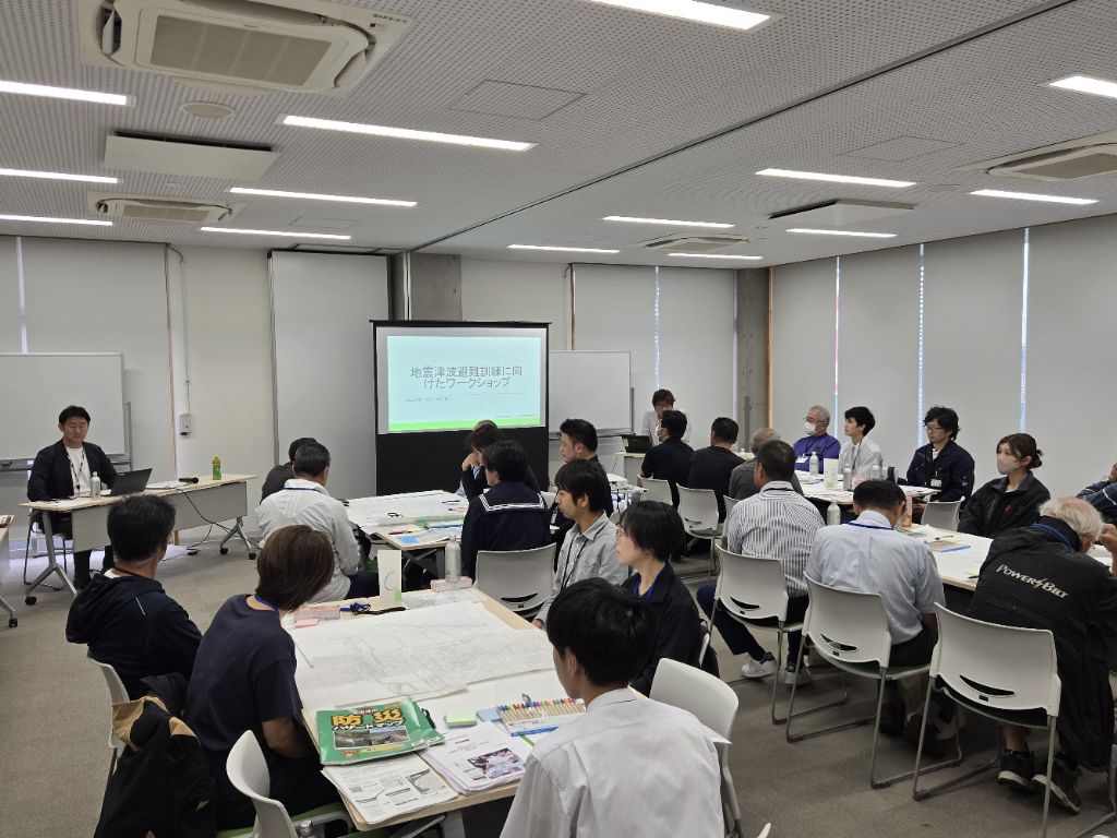 訓練前ワークショップの様子です。参加者が5つのテーブルに分かれて座っており、議論を開始しようとしています。机上には地区全体の白地図や、ハザードマップなどが置いてあります。
