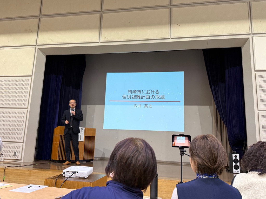 講演台の前の前に立ち、個別避難計画についての資料をスクリーンに投影し、基調講演を行う講師の穴井英之氏です。