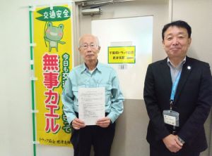 千葉県トラック協会様が実施している、交通安全啓発活動の旗がある事務所の前で、締結した協定書を千葉県トラック協会君津支部桂様（左側）が持って、木更津市総務部前田部参事（右側）と撮影した記念写真。
